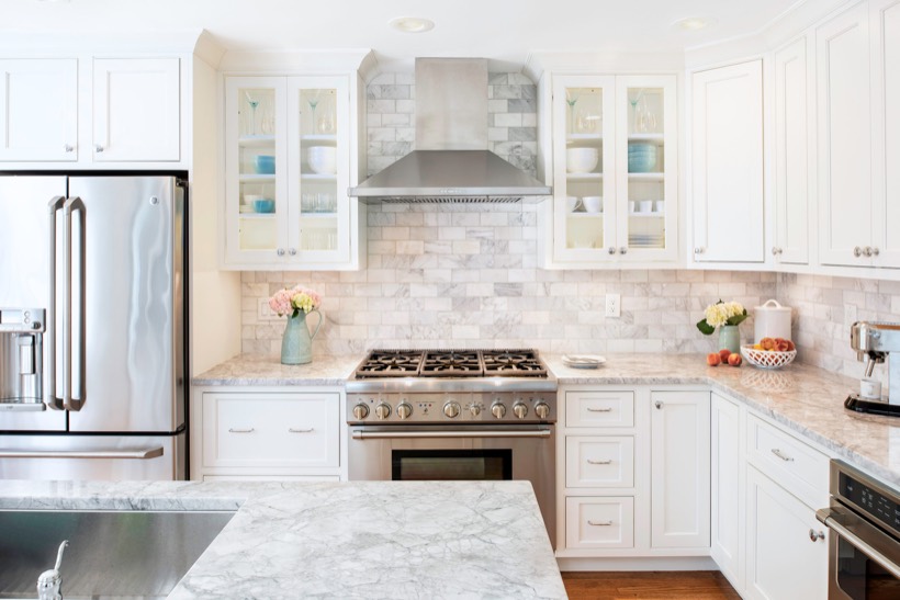 Shiloh cabinetry example, bright, modern kitchen with white kitchen cabinets, stainless steel appliances, marble countertops, and a stylish backsplash. Fresh flowers and bowls decorate the counters, with dishes neatly arranged in glass-front cabinets