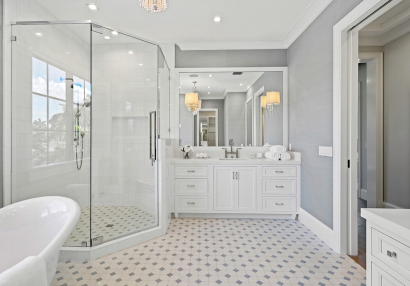 Spacious, modern bathroom with a glass-enclosed shower, sleek Shiloh cabinetry double-sink vanity, freestanding bathtub, geometric tile floor, and elegant lighting fixtures. Walls are light gray with large mirrors above the sinks