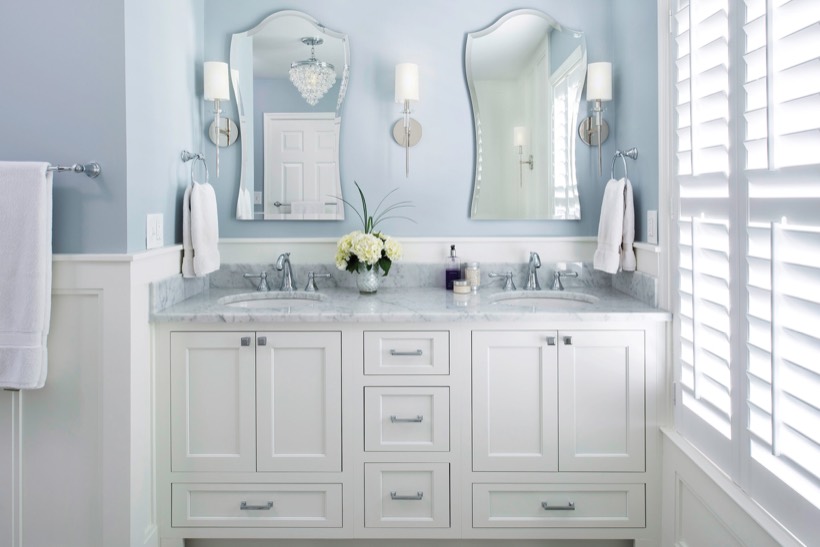 A bright bathroom with a double-sink white vanity from Shiloh cabinetry, marble countertop, two decorative mirrors, wall-mounted sconces, fresh flowers in a vase, and sunlight streaming through white shuttered windows