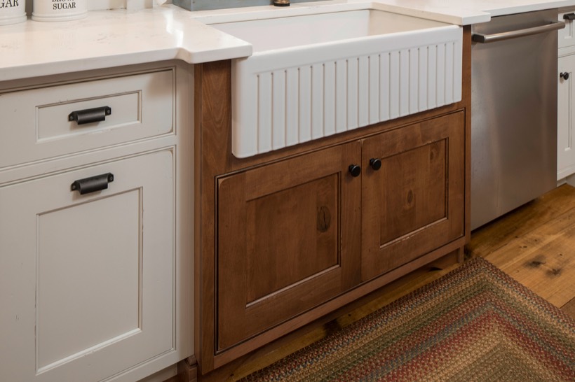 Shiloh cabinetry example, close-up of a farmhouse kitchen sink with a white apron front, set in wooden cabinetry and Kitchen Cabinets, surrounded by white countertops, stainless steel dishwasher, and a woven rug on a wooden floor or DC