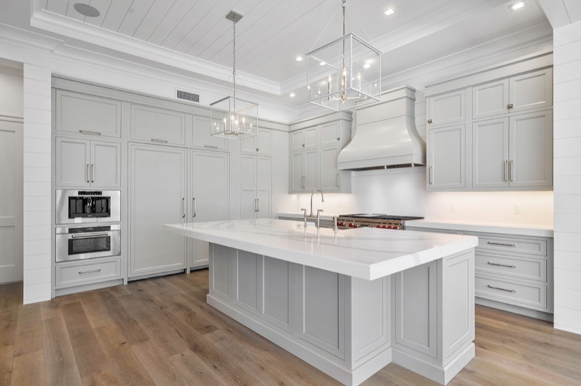 Shiloh cabinetry example, modern kitchen with light gray cabinets, a large white island countertop, stainless steel appliances, built-in oven and microwave, wood flooring, and two rectangular pendant lights hanging above the island