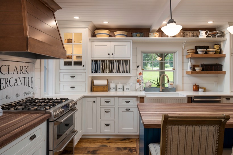 Shiloh cabinetry example, a cozy farmhouse kitchen with white Kitchen Cabinets, open shelves, a gas stove, farmhouse sink, and wood countertops features warm lighting. A window offers a view of the green yard