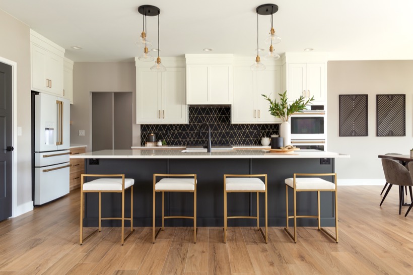 Shiloh cabinetry example, modern kitchen with a large island featuring four gold and white stools, sleek cabinets, black geometric backsplash, pendant lights, built-in ovens, and a vase with green branches