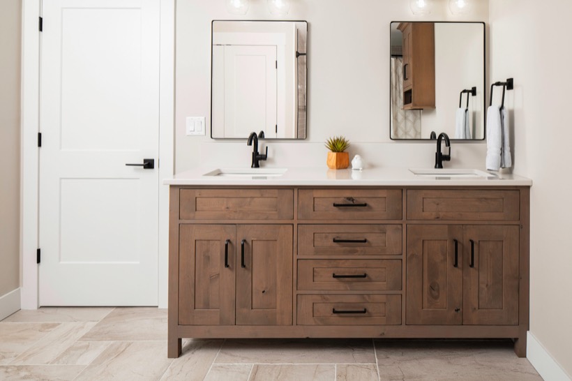 Modern bathroom with a double sink vanity featuring Shiloh cabinetry, black fixtures, two rectangular mirrors, a small potted plant, and white walls and tiles. A white door is on the left side of home