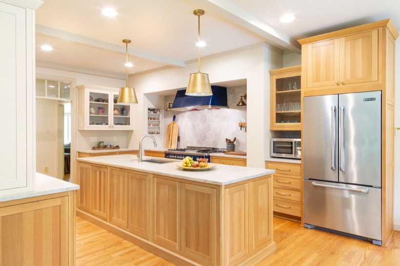 Shiloh cabinetry example, modern kitchen with light wood cabinets, a large island with a sink, stainless steel refrigerator, microwave, fruit bowl, and pendant lights. The backsplash features hexagonal tiles behind the stove