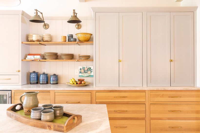 Shiloh cabinetry example, a bright, modern kitchen with light wood drawers, sleek white kitchen cabinets, open shelves displaying pottery and bowls, fruit on the countertop, and a tray with a teapot and cups on the island