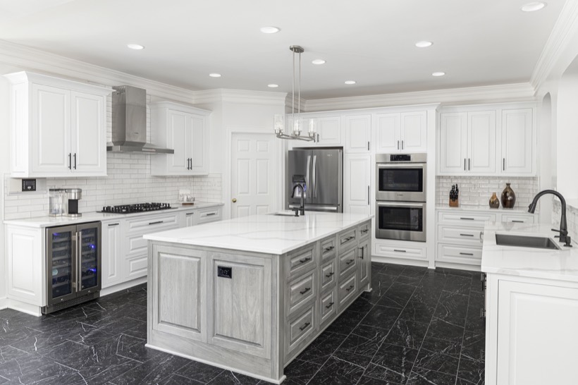 Shiloh cabinetry example, modern kitchen with white kitchen cabinets, stainless steel appliances, marble countertops, a large gray center island, black tile floor, and recessed ceiling lights. A wine cooler, double ovens, and decorative vases are also visible