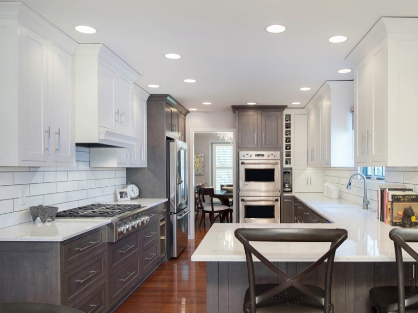 A modern kitchen in with sleek white Shiloh cabinetry above, gray lower cabinets, stainless steel appliances, a white subway tile backsplash, spacious island seating, wooden floors, and ample natural light streaming through the window