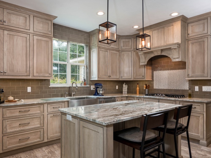 Shiloh cabinetry example, modern MD kitchen with light wood cabinets, granite countertops, a center island with two chairs, stainless steel appliances, double sink under a window, brown tiled backsplash, and two pendant lights hanging from the ceiling