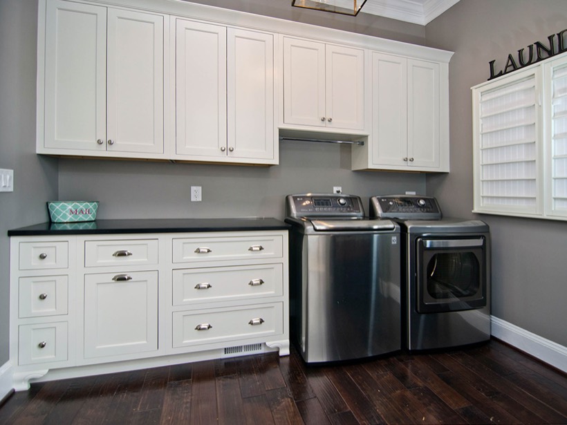 Modern laundry room with dark wood flooring, sleek Shiloh cabinetry in white, a black countertop, and stainless steel washer and dryer. A window with white shutters is on the right wall, and a decorative teal box sits on the counter