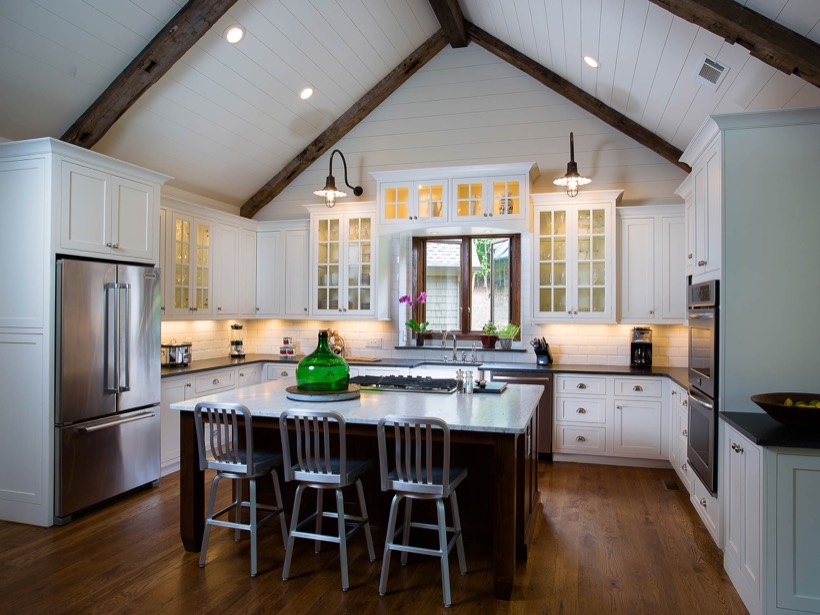Bright, spacious kitchen with vaulted ceiling, exposed wooden beams, sleek Shiloh cabinetry, stainless steel appliances, a large island with three metal barstools, and decorative lighting above. Orchids and a green vase sit on the island