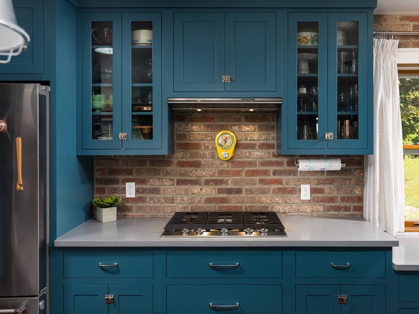Shiloh cabinetry example, a modern MD kitchen features teal cabinets, a brick backsplash, a stainless steel gas stove, a yellow wall clock, glass-front upper kitchen cabinets, and a window with a white curtain to the right