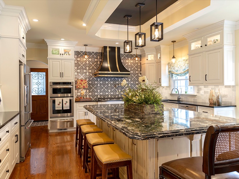 Shiloh cabinetry example, spacious kitchen with white cabinets, patterned tile backsplash, black range hood, marble island countertop, yellow leather bar stools, pendant lights, and hardwood floors. A floral arrangement decorates the island in this stylish home