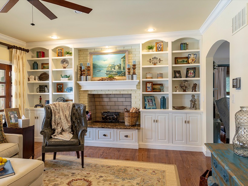 Cozy living room with a decorated fireplace and Shiloh cabinetry built-in shelves filled with books, art, and decor. An armchair, throw blanket, and area rug add warmth; a painting and baskets sit on the mantel. Bright, inviting atmosphere