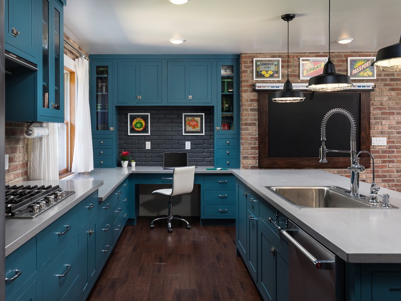 Modern kitchen with teal Shiloh cabinetry, stainless steel appliances, double sink, dark wood floors, and exposed brick walls. A small office nook with a white chair and computer is seamlessly integrated into the cabinetry in home