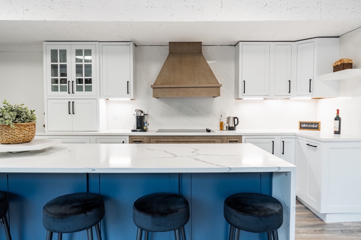 Kitchen display at the Bethesda Cabinets showroom with white cabinets, a wood range hood, a marble island, and blue bar stools.