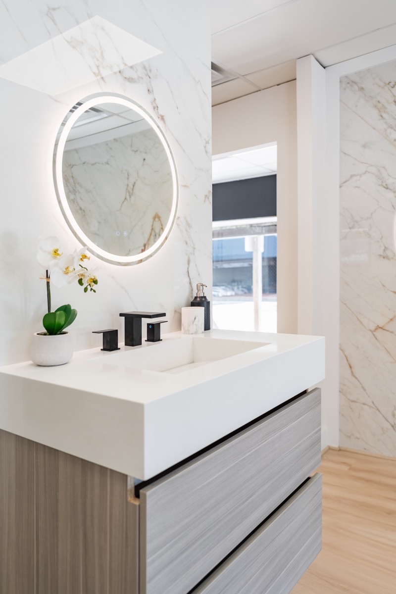 Bathroom vanity display at the Bethesda Cabinets showroom with a white floating vanity, a round mirror, and a marble surround.