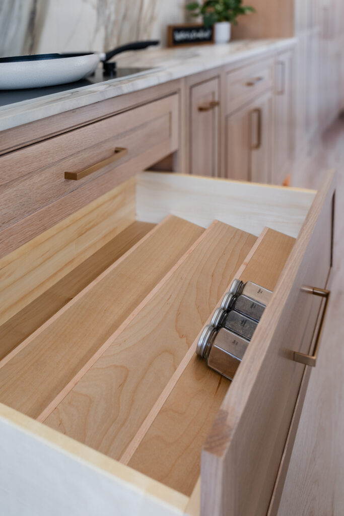 A kitchen drawer with wooden dividers, partially open, holds a few small, labeled spice jars; modern Bethesda Cabinets and a marble countertop are visible in the background.