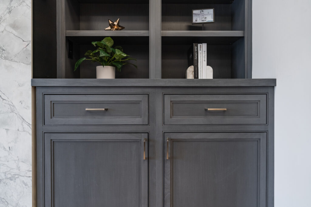 A dark gray Bethesda Cabinet with two drawers and open shelves, holding a potted plant, decorative golden star, small sculpture, and a few books, stands next to a white and gray marble wall in DC or MD.
