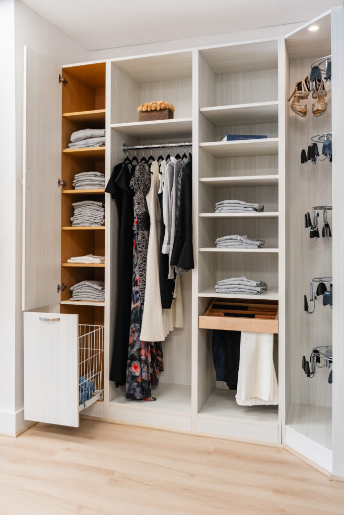 A modern organized closet by Bethesda Cabinets features shelves, hanging clothes including dresses and jackets, neatly folded shirts, shoes on door racks, and a pull-out wire basket—all in a light-colored wooden finish perfect for Maryland and DC homes.
