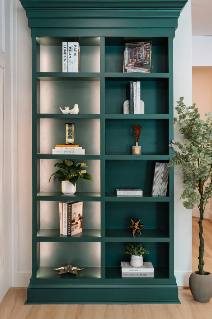A green built-in bookshelf by Bethesda Cabinets displays books, decorative objects, small plants, framed photos, and ornaments. The shelves are neatly arranged, with a potted plant beside the unit on a light wooden Maryland floor.