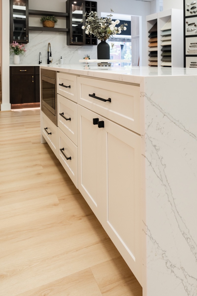 Kitchen display at the Bethesda Cabinets showroom with white shaker cabinets, a marble waterfall island, black hardware, and glass-front upper cabinets.