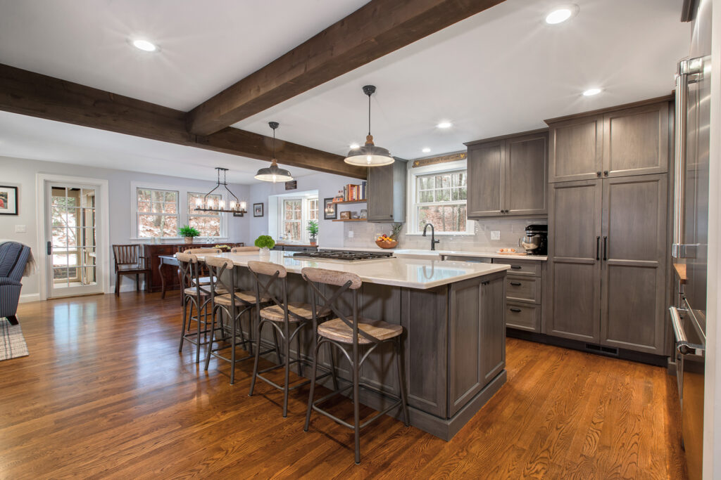 Spacious, modern kitchen with wood flooring, large gray island, four barstools, pendant lights, stainless steel appliances, and exposed wooden ceiling beams. Dining area and windows with natural light in the background.