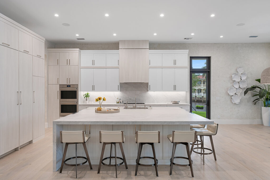 Modern kitchen with light wood cabinets, large marble island with five beige bar stools, built-in ovens, sunflowers in a vase, and a window showing greenery outside. The space is bright with recessed lighting.