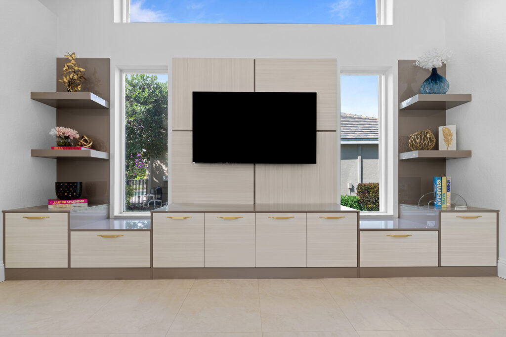 Modern living room with a wall-mounted flat-screen TV centered between two tall windows, built-in cabinets and shelves with decorative items, and bright natural light coming in.