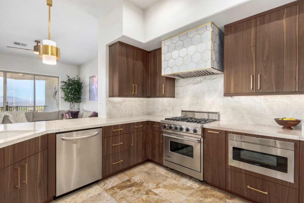 Modern kitchen with dark wood cabinets, stainless steel appliances, marble countertops, and backsplash. A gold-accented hexagonal range hood is above the stove. Open layout leads to a living area with large windows.