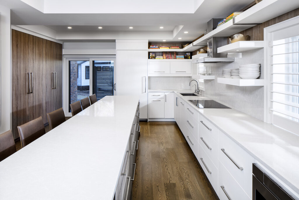 Modern kitchen with white countertops and cabinets, open shelving with dishes, wooden lower cabinets, built-in appliances, and a large island with brown barstools on a wooden floor. Natural light streams in through a window.