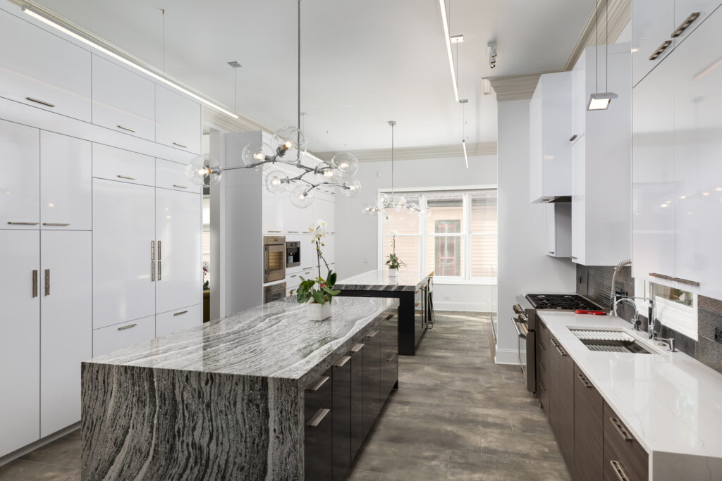 Modern kitchen with two islands featuring gray marble countertops, glossy white cabinets, stainless steel appliances, large windows, pendant lights, and light wood flooring. An orchid plant decorates one island.