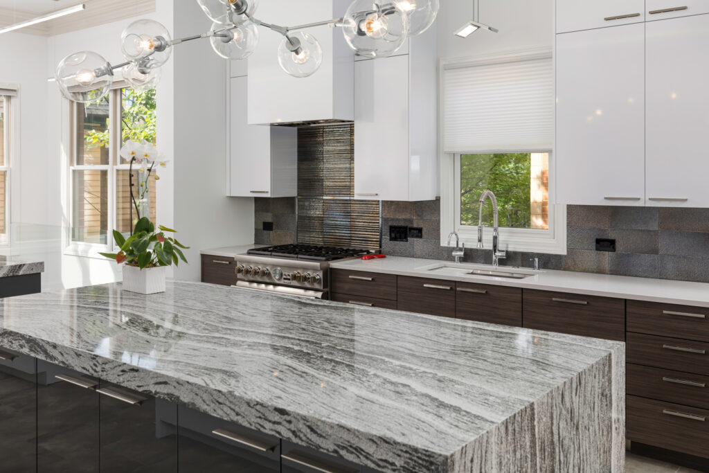 Modern kitchen with a large, gray and white marble island, dark cabinets, white upper cabinets, stainless steel appliances, a plant on the counter, and large windows letting in natural light.