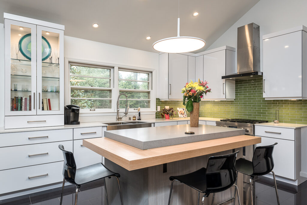 Modern kitchen with white cabinets, green tile backsplash, and stainless steel appliances. An island with black chairs and a concrete countertop features a vase of flowers. A window brings in natural light.