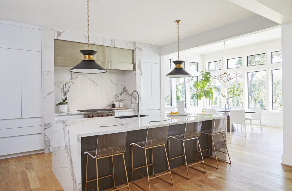 Modern kitchen with white cabinets, marble countertops and backsplash, an island with four clear barstools, gold and black pendant lights, and large windows letting in natural light. Dining area with plants visible in the background.