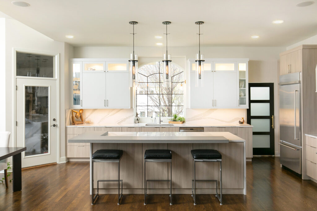 Modern kitchen with white cabinets, marble backsplash, stainless steel appliances, an island with three black stools, pendant lights, and a large window above the sink letting in natural light.