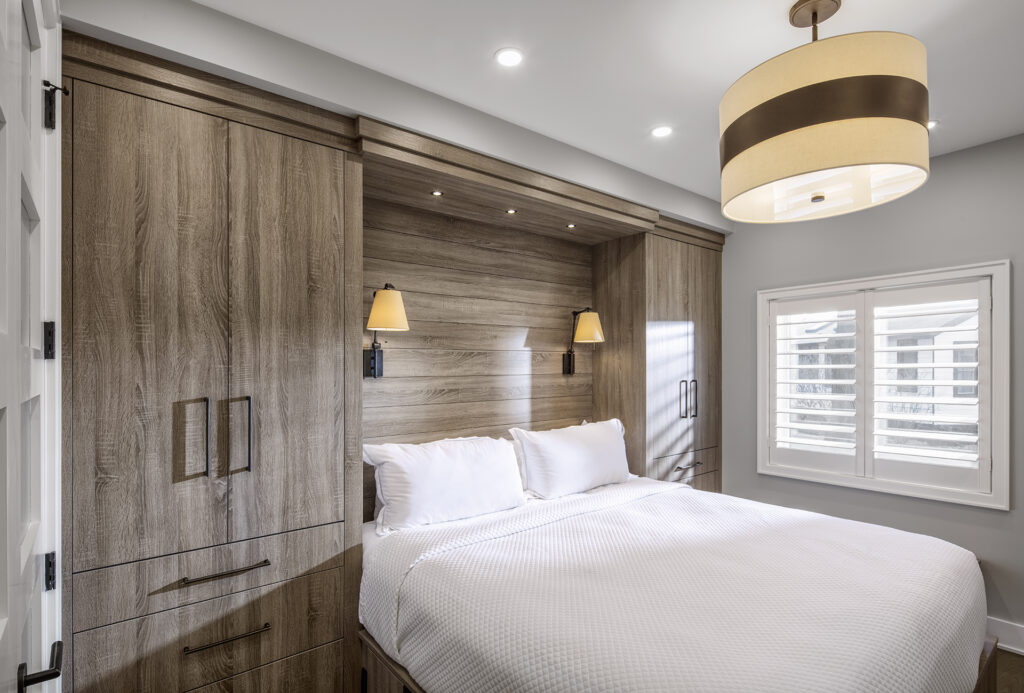 Modern bedroom with wooden built-in cabinets, a double bed with white bedding, two wall-mounted reading lights, a large ceiling lamp, and shuttered windows letting in natural light.