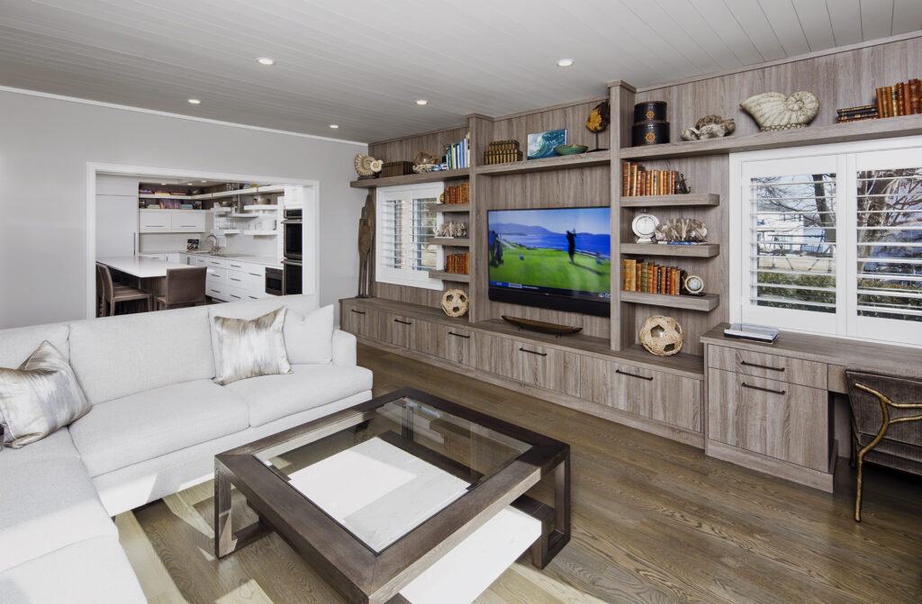 Modern living room with a gray sectional sofa, glass coffee table, built-in wooden shelves with books and decor, a TV displaying a nature scene, and large windows with white shutters; kitchen visible in the background.
