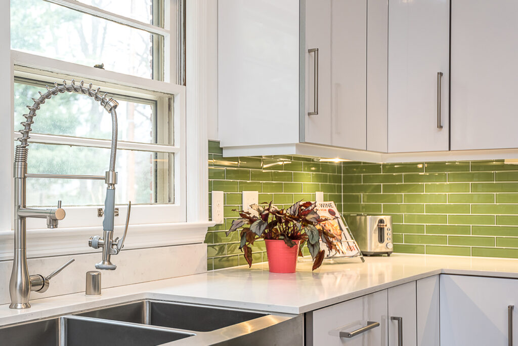 Modern kitchen with white cabinets, green tile backsplash, double stainless steel sink, pull-down faucet, and a potted plant in a red pot on the countertop near a window.