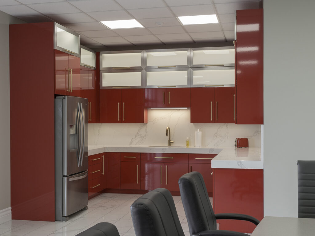 Modern kitchen with glossy red cabinets, stainless steel refrigerator, marble countertops and backsplash, under-cabinet lighting, and a small red toaster. Black chairs are positioned in the foreground.