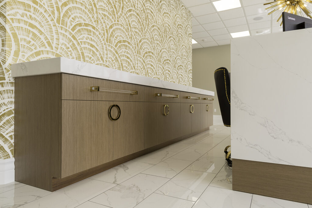 A modern interior features wood cabinets with gold handles, white marble countertops, and a marble-fronted desk. The floor is tiled in white, and the wall has a gold art-deco patterned wallpaper.