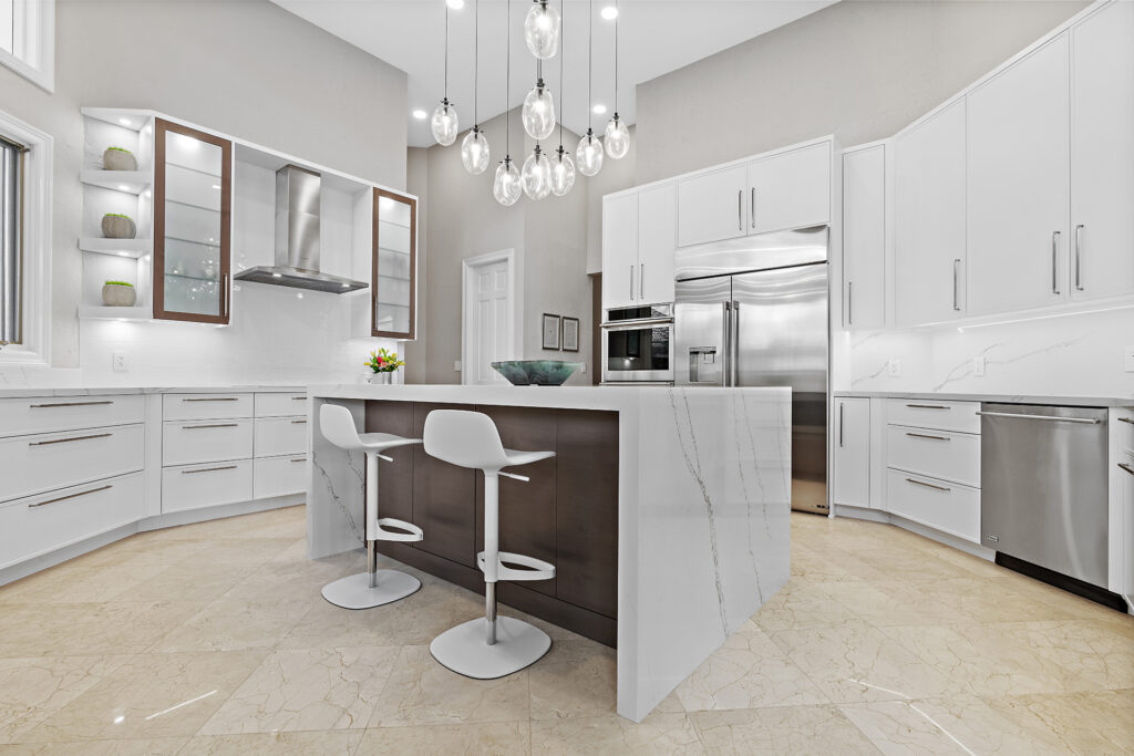 Modern kitchen with white cabinets, marble countertops, stainless steel appliances, an island with two white bar stools, and hanging glass pendant lights. The floor is polished beige tile.