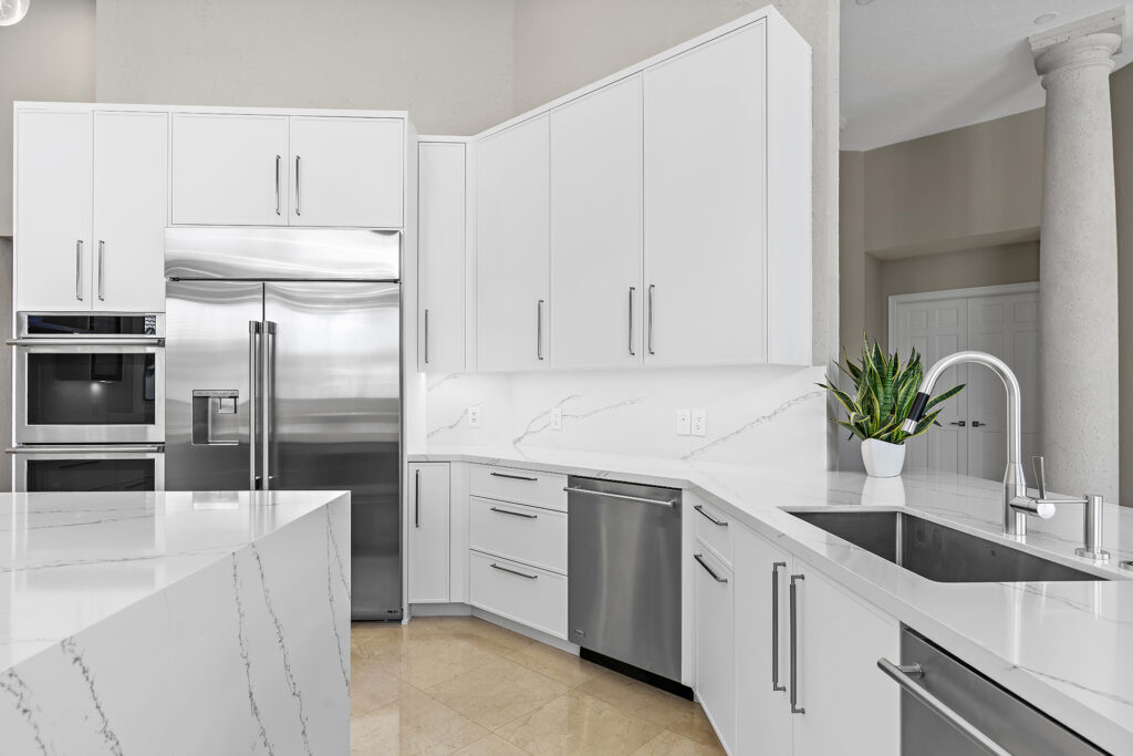 Modern kitchen with white cabinets, stainless steel appliances, marble countertops, built-in oven, double-door fridge, dishwasher, and a sink. A green potted plant sits near the sink for decoration.