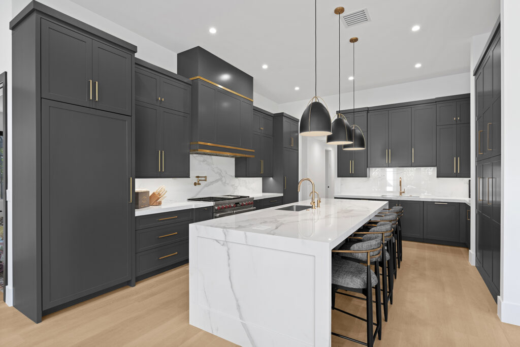 Modern kitchen with dark gray cabinets, gold hardware, white marble countertops and backsplash, large island with bar stools, pendant lights, and light wood flooring. The space is bright and spacious with a minimalist design.