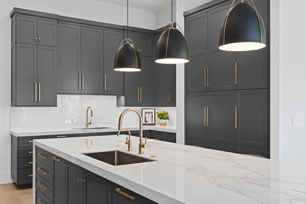 Modern kitchen with dark gray cabinets, white marble countertops, gold hardware, pendant lights, a built-in sink, and minimalist decor, including a small framed photo, a plant, and subtle accents.
