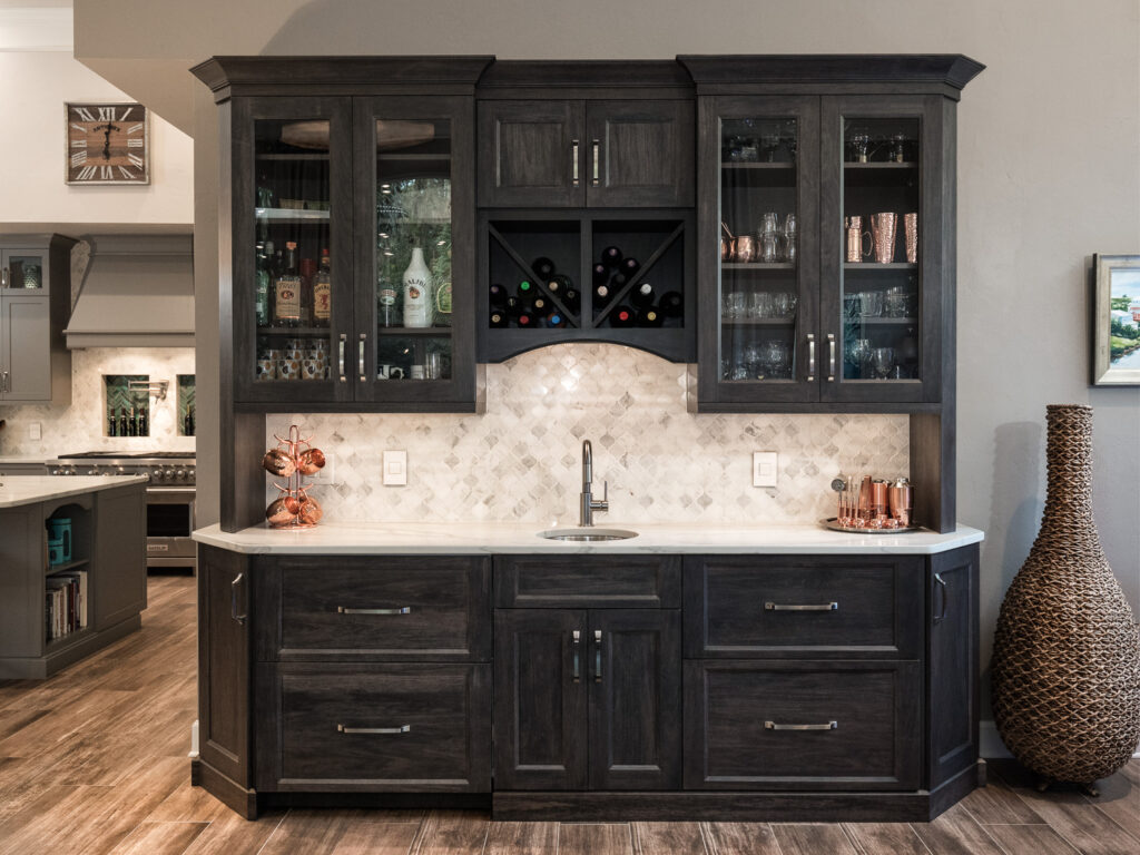 A modern home bar with dark wood cabinets, glass doors displaying glassware, a wine rack, a small sink, and a marble backsplash. Copper bar tools and bottles are on the countertop. A tall woven vase stands to the right.