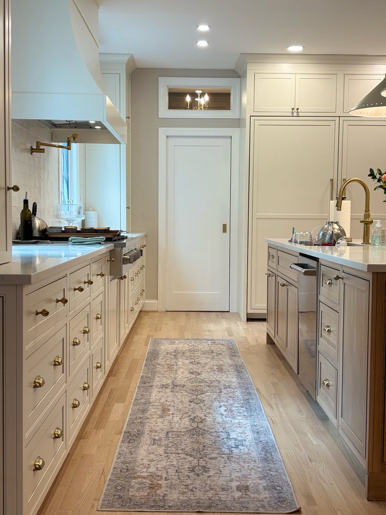 Walkway between Shiloh inset perimeter cabinetry in soft white and a white oak island with MSI quartz countertops, with a runner rug and a pantry door at the end, a Bethesda Cabinets project.