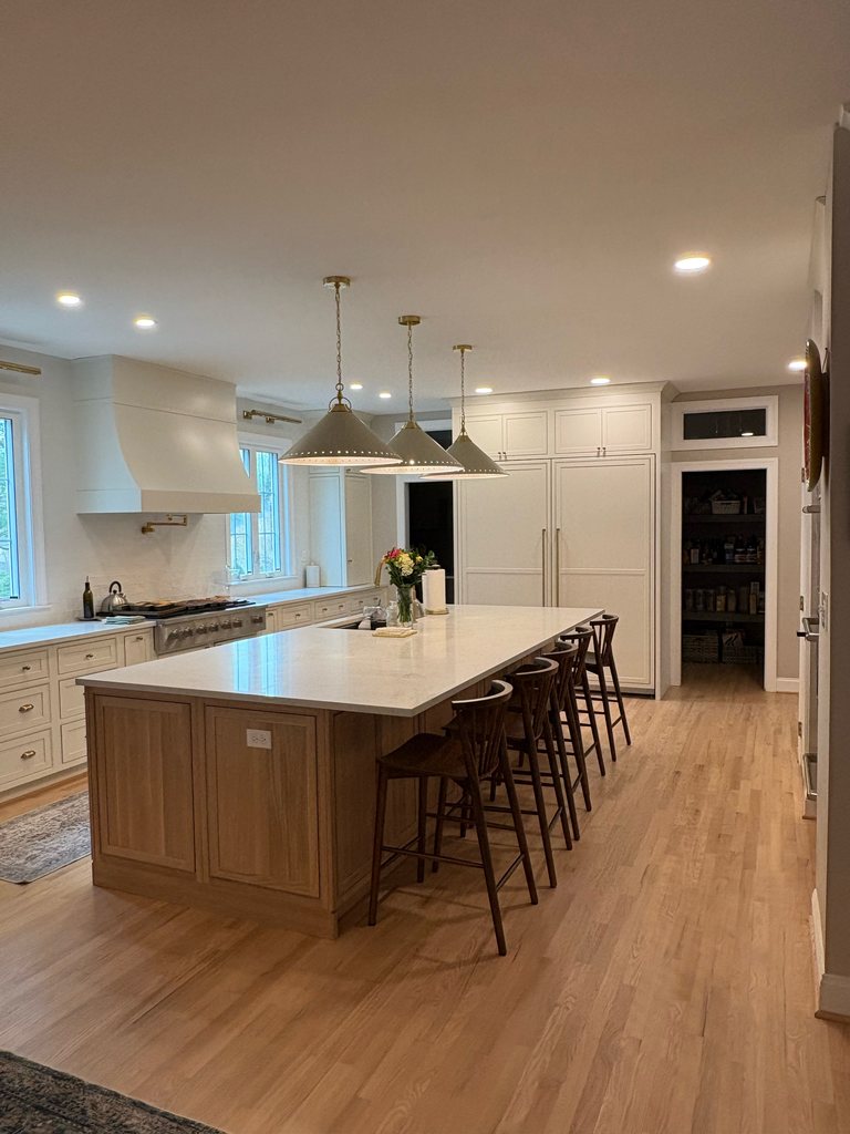 Kitchen with a long white oak island and stools, Shiloh inset cabinetry in soft white, MSI quartz countertops and a range hood, open to the next room, a Bethesda Cabinets project.